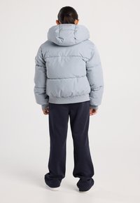 Person mit zurückgebundenem Haar, die eine hellgraue Steppjacke mit Kapuze, schwarze lockere Hosen und weiße Schuhe trägt, steht mit dem Rücken zur Kamera.