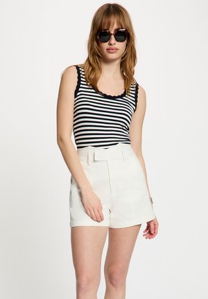 Femme portant un haut rayé noir et blanc sans manches, un short blanc taille haute avec ceinture, et de grandes lunettes de soleil foncées sur un fond uni.