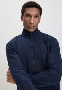 Marineblaues Zip-up-Jackett mit hohem Kragen, das eine glatte Textur und einen taillierten Schnitt aufweist. Der Reißverschluss reicht bis zum Hals, ohne Muster oder Akzente.