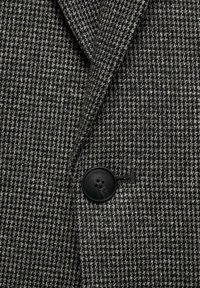 Texturiertes grau-schwarz karierter Tweed-Stoff, der ein Hahnentristmuster zeigt, mit einem schwarzen Knopf, der am Rand sichtbar ist. Glatt und dicht gewebt.