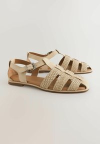 Beige leather sandals with woven pattern on the toe and adjustable ankle strap. Flat sole with minimal hardware accents.
