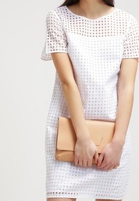 Woman wearing a white short-sleeve perforated dress holding a beige clutch bag with both hands in front of her body.