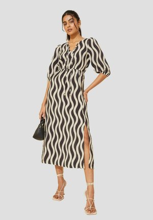 Vestito lungo, nero e beige con motivo a onde, scollatura a V, vita cintrata, maniche corte a palloncino e spacco laterale. Abbinato a sandali con cinturini.