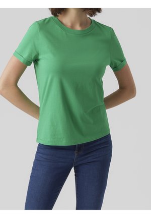 Vrouw met een effen groen T-shirt met korte mouwen en een blauwe spijkerbroek, staand met de handen achter de rug tegen een lichtgrijze achtergrond.