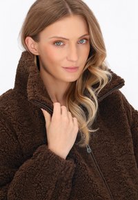 Hnědá fleecová bunda s jemnou strukturou, vysokým límcem a zipovým zapínáním. Model ji nosí, čímž ukazuje hloubku a teplo materiálu.
