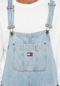 Světle modré džíny na ramínka s kovovými klipsy a přední kapsou se malým logem Tommy Jeans na bílé košili s dlouhým rukávem.