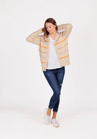 Donna che indossa un cardigan beige con strisce arancioni e viola, una maglietta bianca, jeans blu e sneakers bianche, in piedi con le mani dietro la testa.
