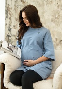 Pregnant woman sitting on beige armchair, wearing blue shirt and black leggings, reading a magazine, with one hand on her belly.
