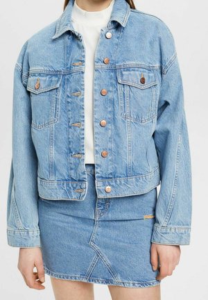 Persona con chaqueta de mezclilla azul claro con botones de cobre y falda de mezclilla a juego, de pie frente a un fondo liso y claro.