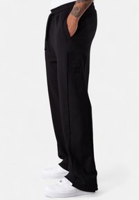 Person in schwarzen Jogginghosen mit seitlicher Applikation und weißen Sneakers, mit einer Hand in der Tasche, vor einem weißen Hintergrund stehend.