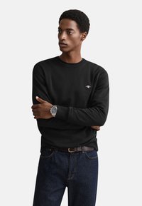 Schwarzer Pullover mit Rundhalsausschnitt aus weichem Stoff, mit kleinem Logo auf der Brust, kombiniert mit dunkelblauen Jeans.