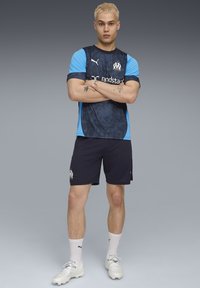 Maillot de sport bleu et noir à manches courtes, présentant un motif géométrique et un logo. Short sombre et chaussures légères, portés avec des chaussettes blanches.