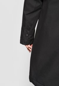 Juodas paltas su keturiais apvaliais juodais mygtukais ir lygiu paviršiumi. Medžiaga atrodo atspari vandeniui ir gerai priglunda.