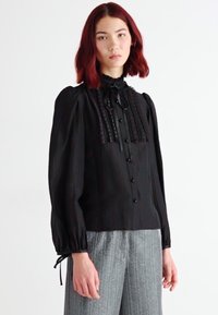Blusa nera con colletto alto a volant, chiusura frontale con bottoni e maniche arricciate. Presenta strisce verticali e un tessuto leggero.