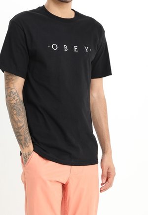 Schwarzes Baumwoll-T-Shirt mit dem weißen Text "OBEY" auf der Brust. Runder Halsausschnitt und kurze Ärmel. Getragen mit korallenfarbenen Shorts.