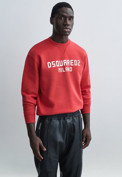 Άνδρας που φοράει κόκκινο φούτερ DSQUARED2 Milano και μαύρο δερμάτινο παντελόνι, στέκεται μπροστά σε απλό γκρι φόντο.