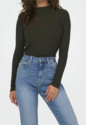 Žena oblečená v černém žebrovaném topu s dlouhými rukávy s vlnitými okraji a světle modrých džínách s vysokým pasem, stojící před jednobarevným pozadím.