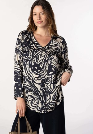 Femme portant un chemisier à manches longues avec un motif floral en noir et blanc, tenant un sac à main marron, une main dans la poche.