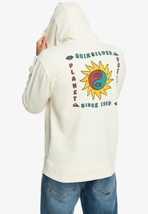Person iført en cremefarvet hættetrøje med en farverig sol og yin-yang grafik og tekst "Quiksilver Planet Positive Since 1969" på ryggen.
