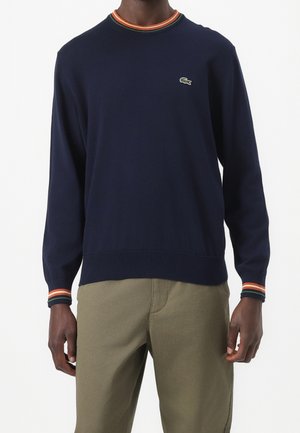 Mann trägt einen marineblauen Pullover mit langen Ärmeln und orange-weißen-grünen gestreiften Bündchen und Kragen, kombiniert mit olivgrünen Hosen.