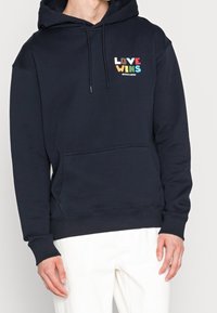 Sudadera azul marino con un bolsillo frontal, capucha con cordón y texto multicolor "EL AMOR GANA" en el pecho. Tejido suave con puños acanalados.