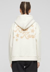 Cream-colored hoodie with beige text and star patterns on the back, featuring a loose fit, hood, and ribbed cuffs. Soft texture.