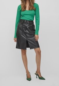 Kvinna iklädd en grön långärmad topp, svart bälte på en fusk läderkjol med slits fram och gröna högklackade slingback-skor mot grå bakgrund.