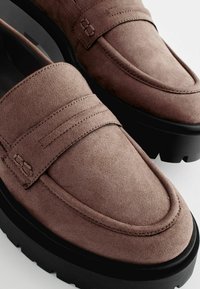 Mocassini in suede marrone con punta arrotondata, cuciture in rilievo e una suola nera spessa con battistrada per la presa. Design minimalista senza hardware visibile.