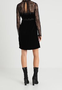 Robe en velours noir avec de longues manches en dentelle, un dos en keyhole, et un détail de taille festonné, associée à des bottes noires à talons jusqu'aux genoux.