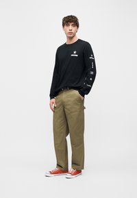 Νεαρός άνδρας που φοράει μαύρο μακρυμάνικο πουκάμισο Converse, ελαι πράσινο παντελόνι cargo και κόκκινα αθλητικά παπούτσια, στέκεται μπροστά σε ένα απλό φωτεινό φόντο.