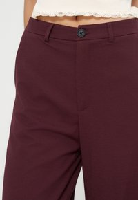 Gros plan sur la taille montrant un pantalon violet foncé avec bouton et fermeture éclair, associé à un top court blanc avec bordure en dentelle.