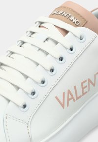 Biela kožená teniska s jemne ružovým koženým prvkom na päte a logom. Má ploché biele šnúrky a kovové očká. Hladká textúra.