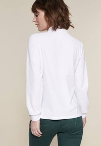 Pullover bianco a maniche lunghe con collo alto e polsini a costine, indossato con pantaloni verdi aderenti con tasche posteriori e dettagli di cucitura.