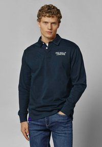 Mornarsko polo majica s dolgimi rokavi, izdelana iz bombaža, z belim logotipom na levi prsni strani. Oblečena z temno modrimi kavbojkami.