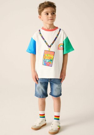 Camiseta blanca con mangas rojas, azules y verdes, que presenta un gráfico de tarjeta de identificación de festival y una impresión arcoíris. Llevada con pantalones cortos de mezclilla y calcetines arcoíris.