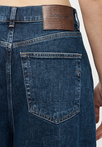 Κοντινή λήψη πίσω τσέπης μπλε τζιν με καφέ υφασμάτινη ετικέτα μάρκας και ορατό πλαϊνό ραφή στη μέση και το χέρι μοντέλου.