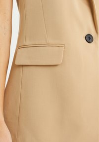 Beige skräddarsydd väst med en strukturerad passform. Har en enkel svart knapp, en klaffficka och strömlinjeformade vertikala sömmar. Slät tygtextur.