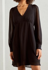 Schwarzes Kleid mit langen, transparenten Ärmeln, tiefem V-Ausschnitt und geraffter Taille. Leichter Stoff, fließende Silhouette und dezenter Glanz.