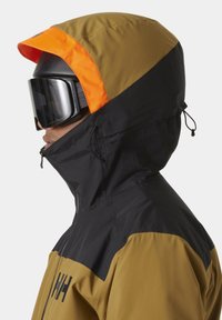 Veste de ski en tissu marron et noir, avec une capuche à visière orange. Les caractéristiques incluent une fermeture éclair, des cordons de serrage et de grosses lunettes de ski.
