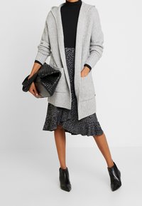 Light gray knitted cardigan with a hood, ribbed texture, and pockets, paired with a black leopard print dress and ankle boots.