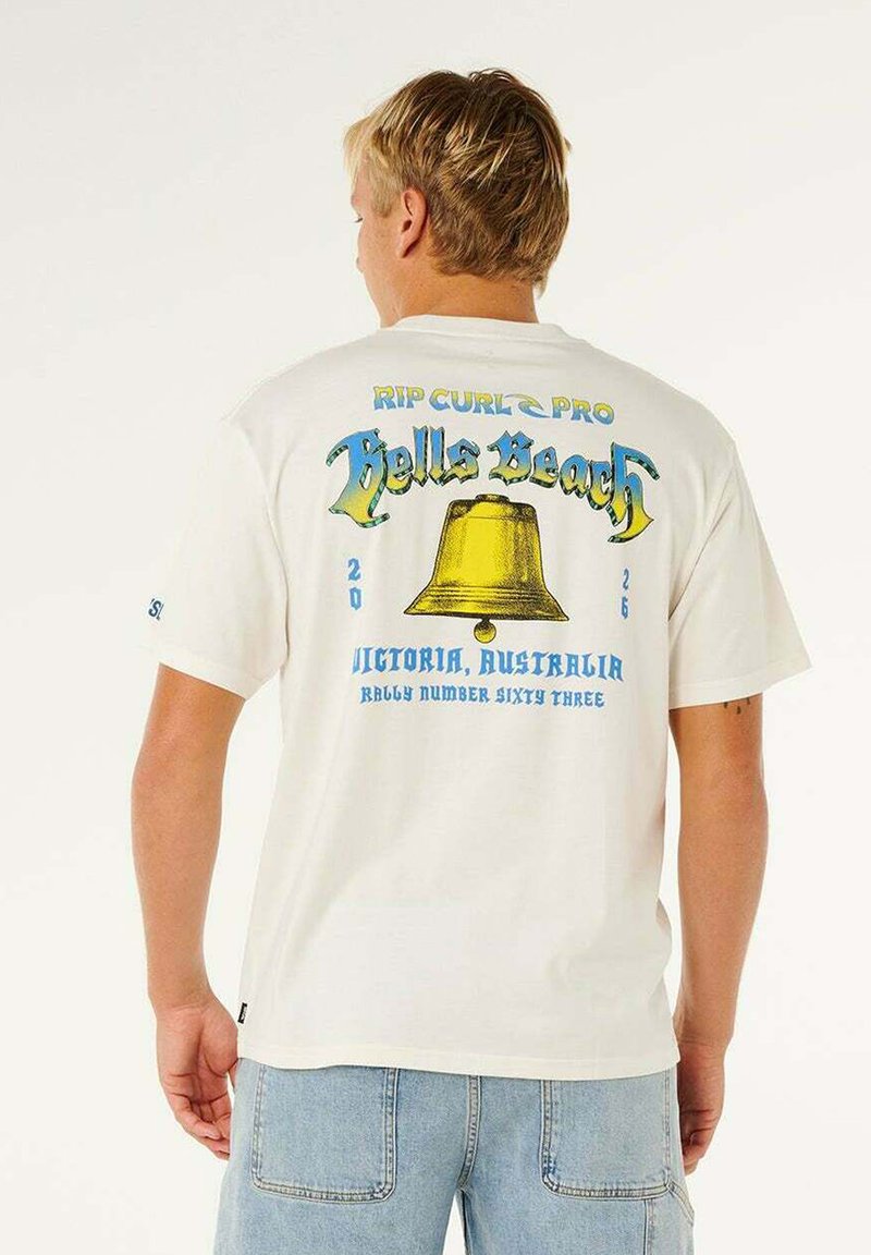 Jeune homme aux cheveux blonds courts portant un t-shirt blanc avec une cloche jaune et le texte "Bells Beach, Victoria, Australia" au dos.