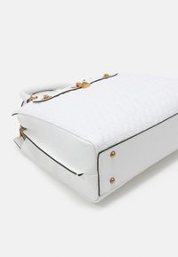 Bílá prošívaná kabelka se zlatými kovovými doplňky, plochým dnem a černým lemováním podél okrajů. Texturovaný povrch se strukturovaným designem.