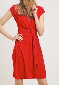 Robe rouge avec des manches courtes, devant drapé et une finition torsadée à la taille. Tissu lisse avec une longueur au genou.