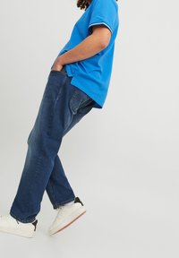 Person wearing blue polo shirt, dark blue jeans, and white sneakers, standing with hands in pockets against a plain white background.
