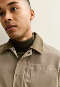 Beżowa kurtka z kołnierzem z sztruksu, srebrnym zatrzaskiem i kieszenią na piersi, noszona na ciemnozielonym swetrze z przędzy typu ribbed.