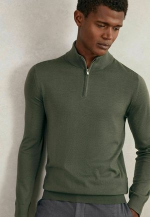 Uomo che indossa un maglione con cerniera a maniche lunghe di colore verde oliva, in piedi contro un muro grigio chiaro, che guarda verso il basso.