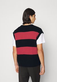 Gilet sans manches en tricot featuring des rayures horizontales rouges et navy. Encolure ronde, texture côtelée et coupe ample à l'arrière.