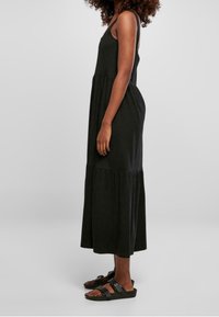 Kvinna som står sidledes iklädd en ärmlös svart midikjol och svarta sandaler med spänne mot en enkel ljus bakgrund.