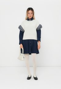 Elfenbeinfarbener Strickpullover mit rüschengesäumten, gestreiften Ärmeln, über einem marineblauen, plissierten Kleid getragen. Das Model hält eine kleine weiße Handtasche und trägt dazu weiße Strumpfhosen und schwarze Ballerinas.