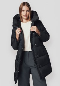 Czarna puchowa kurtka z dużym kapturem, z pikowanym wzorem i srebrnymi zatrzaskami, noszona na lekki sweter i szare spodnie.